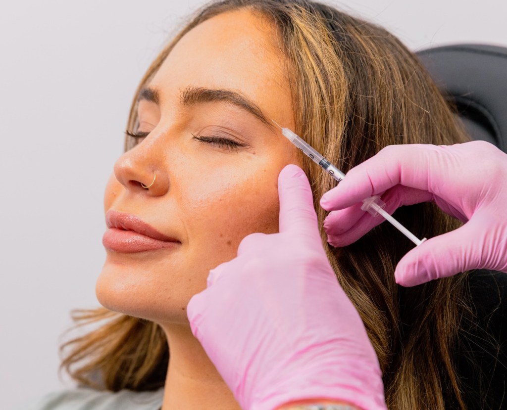 A Skin Boutique provider's hands hold a hypodermic pointed at the brow area of a beautiful female patient. The patient is using dermal fillers in Oakland County, MI, to balance their facial features.